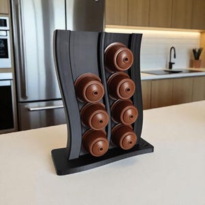 Puede incluir: Un soporte vertical negro para c&aacute;psulas de caf&eacute; con dise&ntilde;o ondulado, que cuenta con dos columnas para un total de ocho c&aacute;psulas de caf&eacute; marrones. El soporte tiene una base rectangular plana y presenta un acabado mate y liso en toda su estructura.