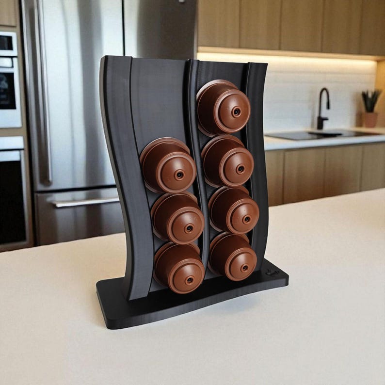 Puede incluir: Un soporte vertical negro para c&aacute;psulas de caf&eacute; con dise&ntilde;o ondulado, que cuenta con dos columnas para un total de ocho c&aacute;psulas de caf&eacute; marrones. El soporte tiene una base rectangular plana y presenta un acabado mate y liso en toda su estructura.