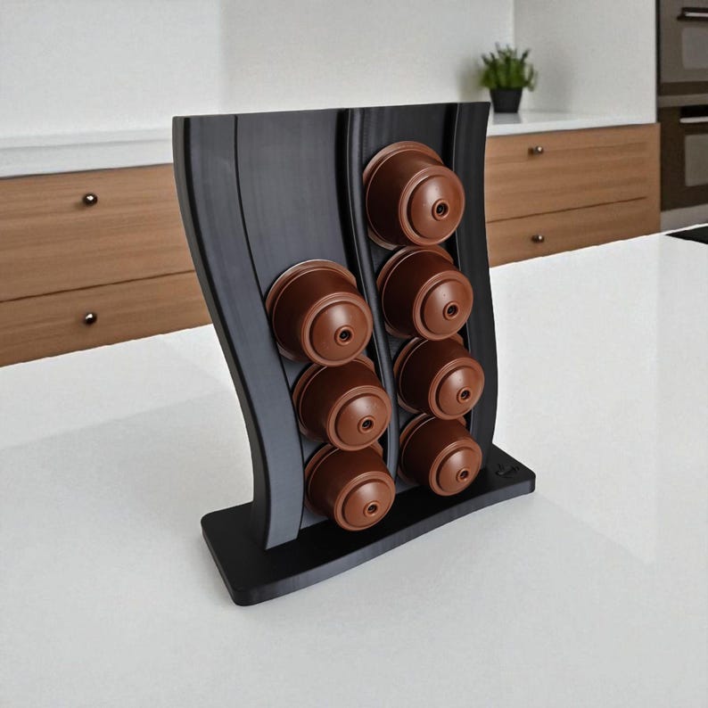 Puede incluir: Un soporte vertical negro para c&aacute;psulas de caf&eacute; con dise&ntilde;o curvo, que cuenta con dos columnas para un total de ocho c&aacute;psulas marrones. El soporte tiene una base ancha y plana, con un acabado mate liso para colocar sobre la encimera.