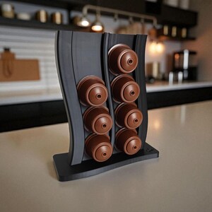 Puede incluir: Un soporte vertical negro para c&aacute;psulas de caf&eacute; con dise&ntilde;o ondulado, que cuenta con dos columnas para un total de ocho c&aacute;psulas de caf&eacute; marrones. El soporte tiene una base rectangular y un acabado liso y mate para almacenamiento.