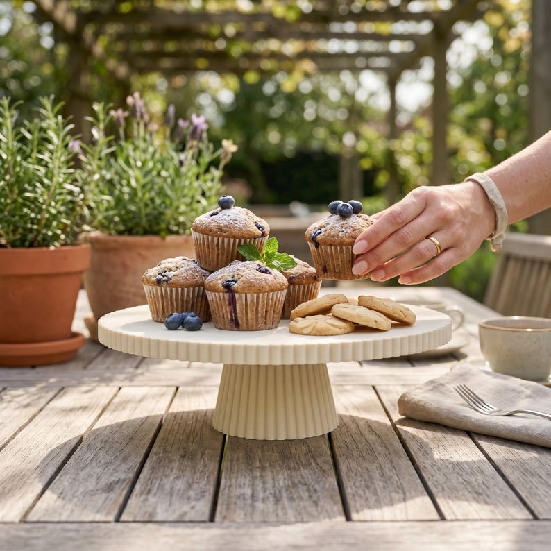 May include: A cream-colored, fluted ceramic cake stand holds several blueberry muffins in paper liners. The muffins are topped with fresh blueberries and a sprig of mint. A few round cookies are arranged on the edge of the stand.
