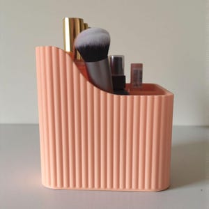 May include: A peach-coloured plastic desk organiser with a vertical ribbed texture. The container features a sloped top edge and holds several items, including a makeup brush with white bristles, a gold-capped bottle, and a small spray bottle.