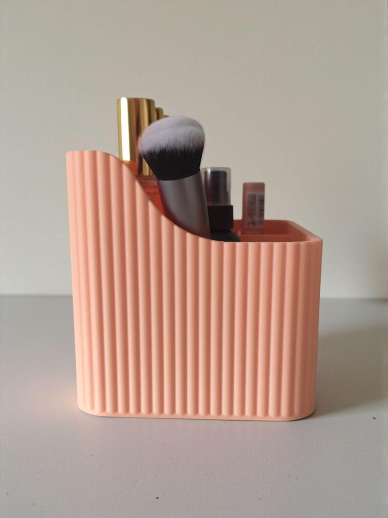 May include: A peach-coloured plastic desk organiser with a vertical ribbed texture. The container features a sloped top edge and holds several items, including a makeup brush with white bristles, a gold-capped bottle, and a small spray bottle.