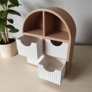 May include: A tan arched desktop organizer featuring four compartments. Two upper sections are open, while two lower sections contain white drawers. The bottom drawer has a vertical ribbed texture on its front face, and both drawers feature a circular cutout.