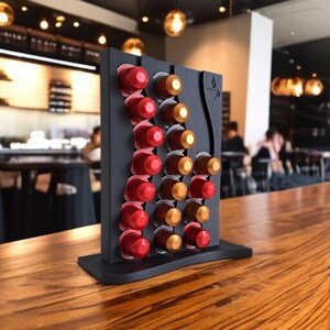 May include: A black vertical coffee pod holder with a wavy design, featuring three columns of slots filled with red and gold-toned coffee capsules. The stand has a flat base and a small coffee cup icon engraved on the upper right corner.