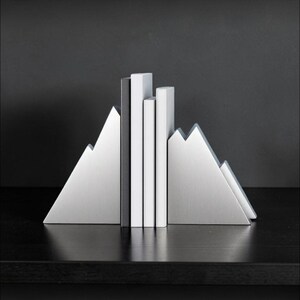 Peut inclure: Une paire de serre-livres en m&eacute;tal argent&eacute; en forme de sommets de montagne, maintenant trois livres verticaux aux dos blancs et noirs. Les serre-livres pr&eacute;sentent une finition m&eacute;tallique bross&eacute;e et une silhouette triangulaire dentel&eacute;e.