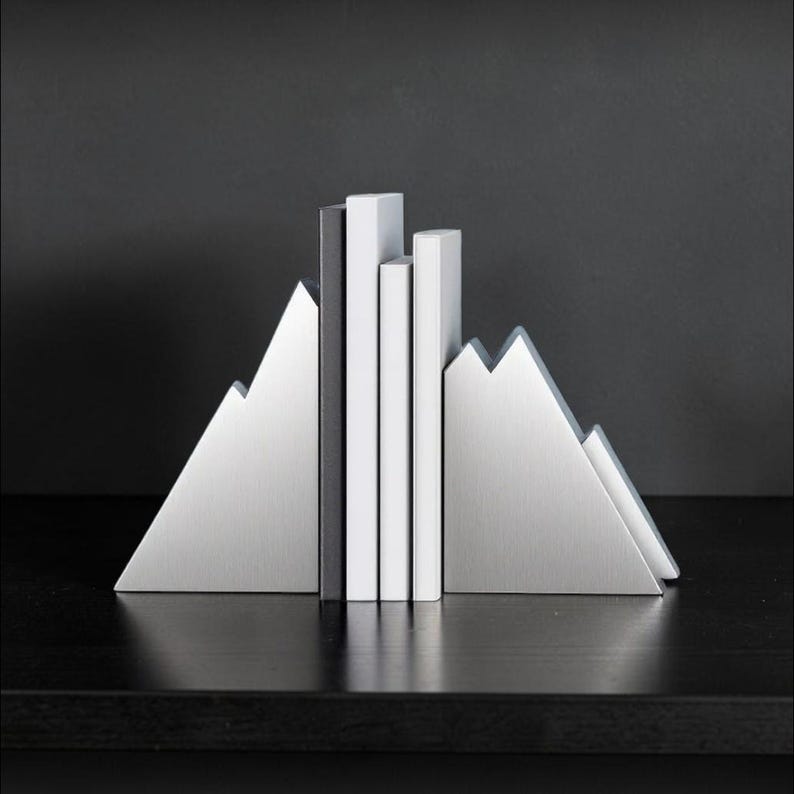 Peut inclure: Une paire de serre-livres en m&eacute;tal argent&eacute; en forme de sommets de montagne, maintenant trois livres verticaux aux dos blancs et noirs. Les serre-livres pr&eacute;sentent une finition m&eacute;tallique bross&eacute;e et une silhouette triangulaire dentel&eacute;e.