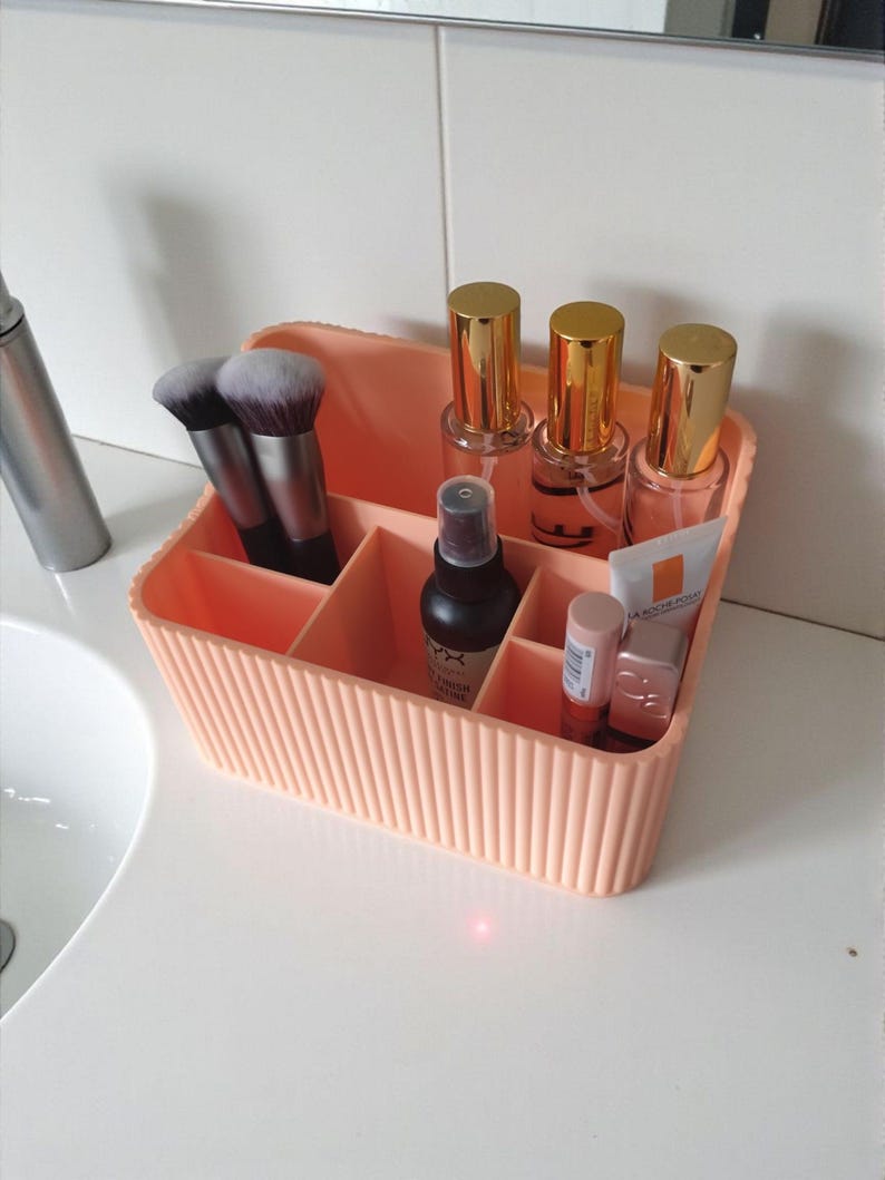 May include: A peach-coloured plastic makeup organiser with vertical ribbed detailing and multiple compartments. It contains two makeup brushes, three glass bottles with gold caps, a black spray bottle, and two small tubes of skincare products.