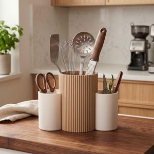 May include: A three-piece kitchen utensil holder set featuring a tall, ribbed, light brown central cylinder flanked by two smaller, smooth, off-white cylindrical containers. The set holds various metal kitchen tools, including a whisk, spatula, and grater.