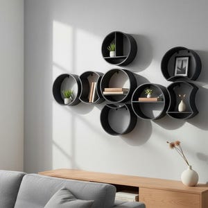 May include: A set of seven interconnected black circular wall shelves arranged in a cluster. The shelves feature various internal dividers and horizontal surfaces, holding small potted plants, books, and a framed picture, mounted on a light gray wall.