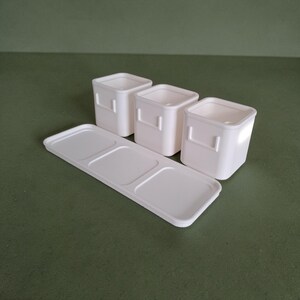 May include: A set of three off-white square containers with small side handles, arranged behind a matching rectangular tray featuring three recessed square sections designed to hold the containers securely in place on a flat surface.
