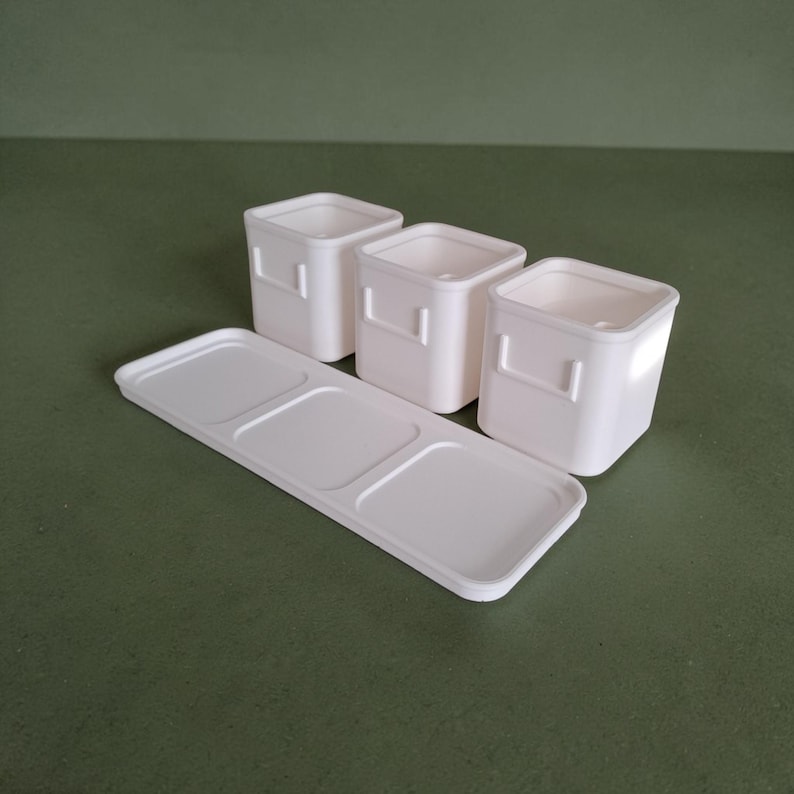 May include: A set of three off-white square containers with small side handles, arranged behind a matching rectangular tray featuring three recessed square sections designed to hold the containers securely in place on a flat surface.