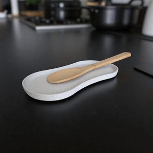 May include: An oval-shaped, speckled light grey spoon rest with a slightly curved, irregular edge. A light-coloured wooden spoon with a smooth, rounded bowl and a long, straight handle rests diagonally across the centre of the spoon rest.