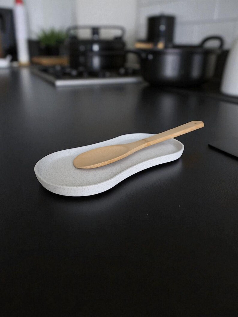 May include: An oval-shaped, speckled light grey spoon rest with a slightly curved, irregular edge. A light-coloured wooden spoon with a smooth, rounded bowl and a long, straight handle rests diagonally across the centre of the spoon rest.