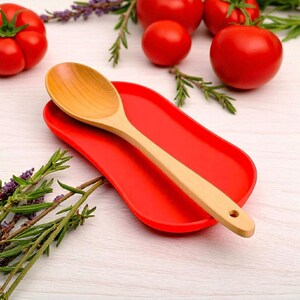 May include: A light-coloured wooden spoon with a rounded bowl and a handle featuring a small circular hole at the end, resting diagonally across a smooth, oval-shaped, solid red ceramic spoon rest with slightly raised edges.