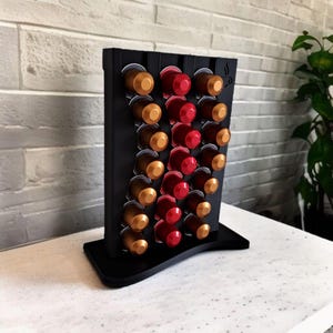 May include: A black vertical coffee pod holder with a wavy design, featuring three columns of slots filled with metallic gold and red coffee capsules. The stand has a flat base and a small coffee cup icon engraved in the top right corner.