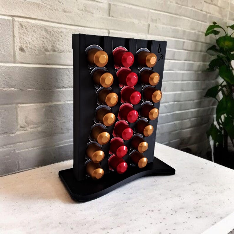 May include: A black vertical coffee pod holder with a wavy design, featuring three columns of slots filled with metallic gold and red coffee capsules. The stand has a flat base and a small coffee cup icon engraved in the top right corner.