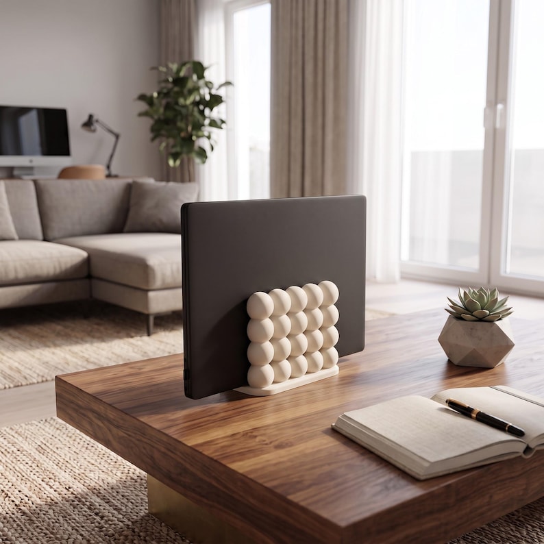 May include: A dark gray laptop held upright by a cream-colored stand featuring a grid of spherical, ball-like shapes. The stand has a flat base and supports the laptop vertically, showcasing a textured, rounded design on its front-facing surface.