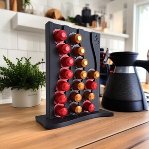 May include: A matte black vertical coffee pod holder with a wavy design, featuring rows of red and gold coffee capsules. The stand has a flat base and a small coffee cup icon engraved on the upper right side, holding multiple pods securely.