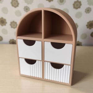 May include: A small, arched, light brown storage organizer featuring two open top compartments and four drawers. The two top drawers are smooth white, while the two bottom drawers feature a vertical ribbed texture, each with a semi-circular cutout.