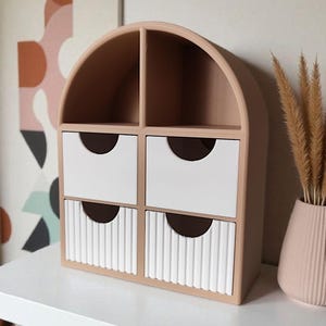 May include: A beige arched desktop organizer featuring two open top compartments and four drawers. The top two drawers have smooth white fronts, while the bottom two drawers feature a vertical ribbed texture. Each drawer has a semi-circular cutout.
