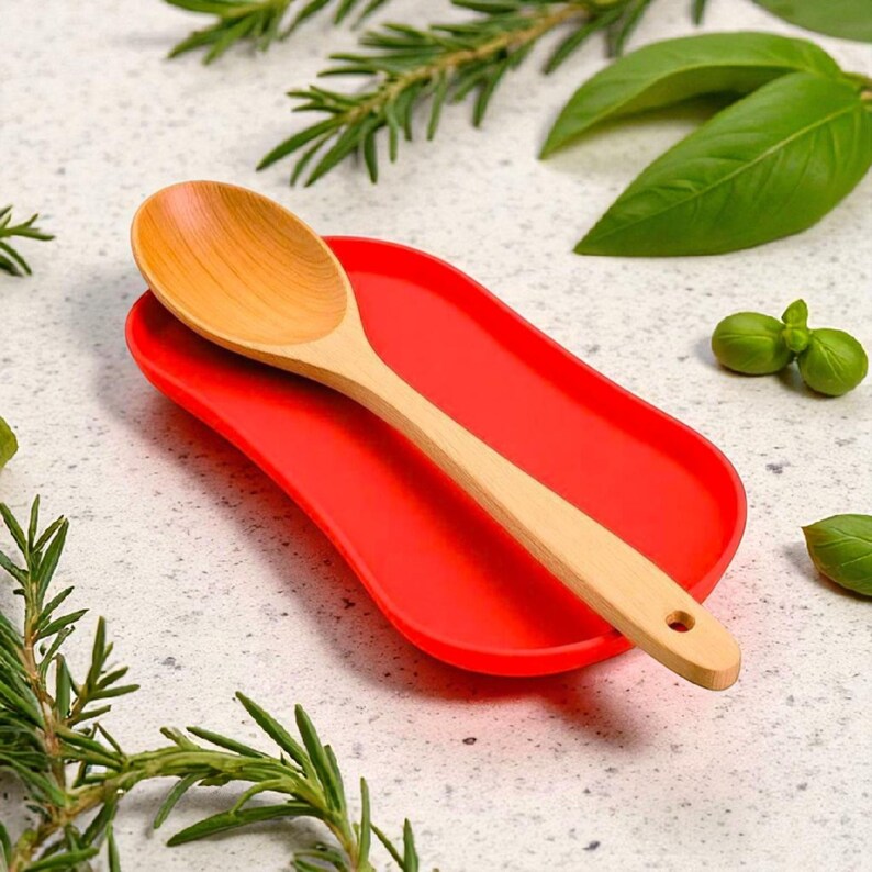 May include: A light-coloured wooden spoon with a rounded bowl and a handle featuring a small circular hole at the end, resting diagonally across a bright red, oval-shaped silicone spoon rest with a smooth, slightly concave surface.