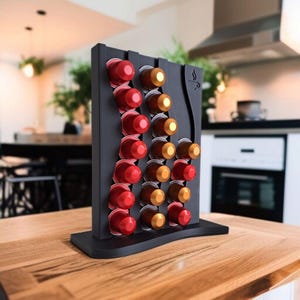 May include: A black vertical coffee pod holder with a wavy design, featuring three columns of slots filled with red and gold coffee capsules. The stand has a flat base and a small coffee cup icon engraved on the upper right side of the frame.