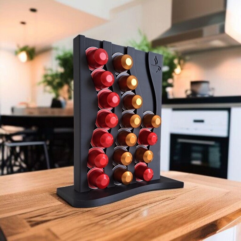 May include: A black vertical coffee pod holder with a wavy design, featuring three columns of slots filled with red and gold coffee capsules. The stand has a flat base and a small coffee cup icon engraved on the upper right side of the frame.