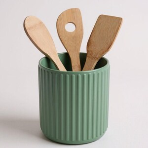 May include: A sage green cylindrical utensil holder with a vertical ribbed texture containing three light brown wooden kitchen utensils, including a rounded spoon, a slotted turner with a circular hole, and a flat-edged spatula.