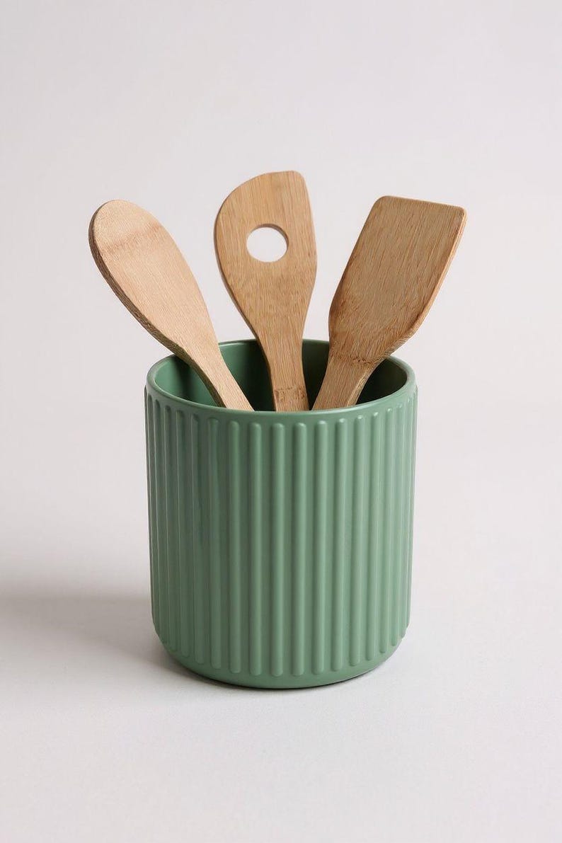 May include: A sage green cylindrical utensil holder with a vertical ribbed texture containing three light brown wooden kitchen utensils, including a rounded spoon, a slotted turner with a circular hole, and a flat-edged spatula.