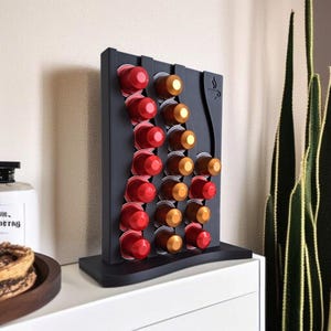 May include: A black vertical coffee pod holder with a wavy design, featuring three columns of slots filled with red and gold coffee capsules. The top right corner of the holder displays a small engraved icon of a steaming coffee cup.