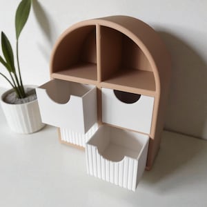 May include: A tan, arch-shaped desktop organizer featuring two open upper compartments and two lower drawers. The drawers have white fronts, with the bottom drawer featuring a vertical ribbed texture and each drawer having a semi-circular cutout handle.