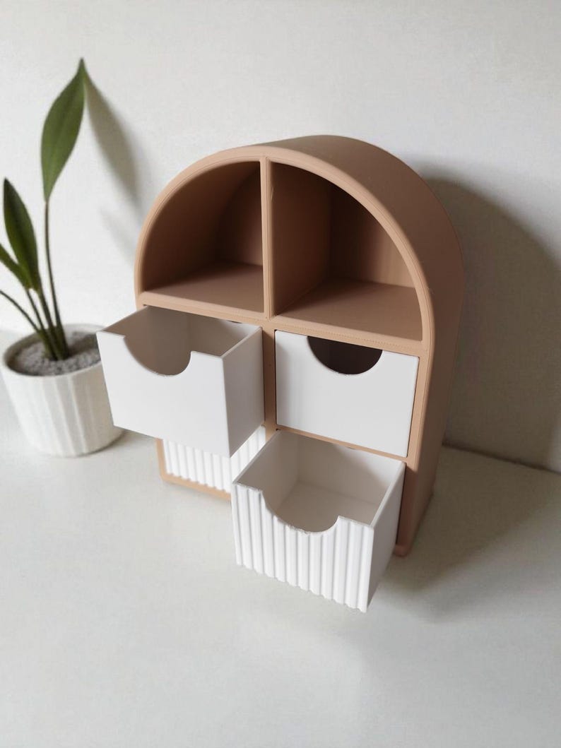 May include: A tan, arch-shaped desktop organizer featuring two open upper compartments and two lower drawers. The drawers have white fronts, with the bottom drawer featuring a vertical ribbed texture and each drawer having a semi-circular cutout handle.