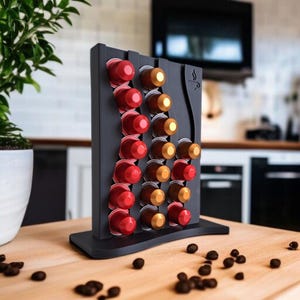 May include: A matte black vertical coffee pod holder with a wavy design, featuring three columns of slots filled with red and gold coffee capsules. The stand has a flat base and a small coffee cup icon engraved on the upper right side.