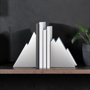Peut inclure: Une paire de serre-livres en m&eacute;tal argent&eacute; en forme de sommets de montagne, maintenant trois livres verticaux aux dos blancs. Les serre-livres pr&eacute;sentent une finition bross&eacute;e et une silhouette triangulaire, soutenant les livres sur une &eacute;tag&egrave;re.