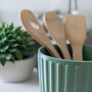 May include: A sage green cylindrical utensil holder with vertical ribbed detailing containing three light-colored wooden kitchen utensils, including a slotted spoon, a solid spoon, and a flat-edged spatula, arranged upright inside.