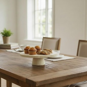 May include: A round, off-white ceramic cake stand with a fluted, ribbed pedestal base and a matching fluted rim. The stand is positioned on a wooden table and holds three golden-brown muffins and two chocolate chip cookies on its flat surface.