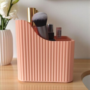 May include: A peach-coloured plastic desk organiser with a vertical ribbed texture and a sloped top edge. The container holds various cosmetic items, including a makeup brush with black and white bristles, a gold-capped bottle, and small tubes.
