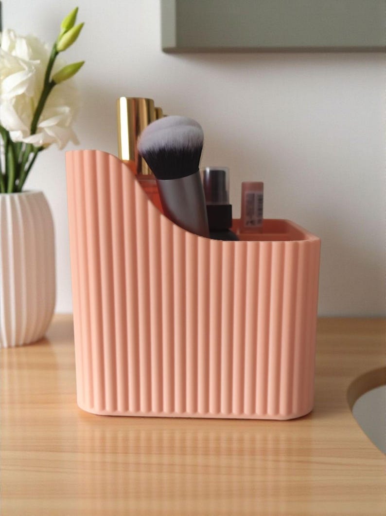 May include: A peach-coloured plastic desk organiser with a vertical ribbed texture and a sloped top edge. The container holds various cosmetic items, including a makeup brush with black and white bristles, a gold-capped bottle, and small tubes.