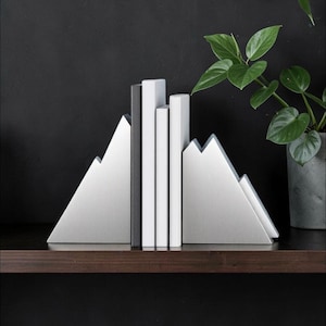 Peut inclure: Une paire de serre-livres en m&eacute;tal argent&eacute; en forme de sommets de montagne, maintenant trois livres aux dos blanc et noir. Les serre-livres pr&eacute;sentent une finition bross&eacute;e lisse et une silhouette triangulaire, soutenant les livres verticaux.