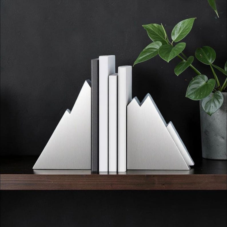 Peut inclure: Une paire de serre-livres en m&eacute;tal argent&eacute; en forme de sommets de montagne, maintenant trois livres aux dos blanc et noir. Les serre-livres pr&eacute;sentent une finition bross&eacute;e lisse et une silhouette triangulaire, soutenant les livres verticaux.