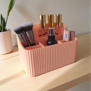 May include: A peach-coloured rectangular plastic desk organiser with vertical ribbed detailing. The organiser features multiple compartments containing two makeup brushes, three glass bottles with gold caps, a black spray bottle, and two lipsticks.
