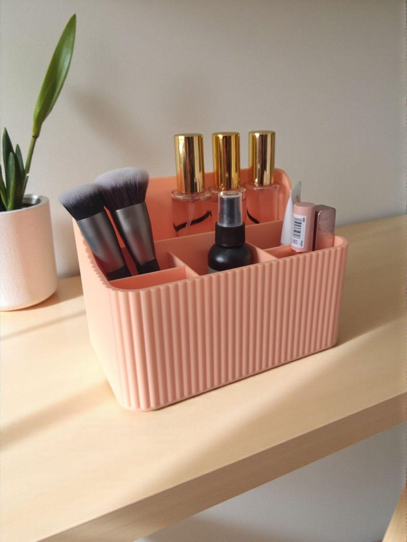 May include: A peach-coloured rectangular plastic desk organiser with vertical ribbed detailing. The organiser features multiple compartments containing two makeup brushes, three glass bottles with gold caps, a black spray bottle, and two lipsticks.