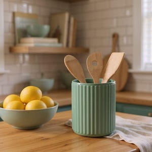 May include: A sage green cylindrical utensil holder with vertical ribbed detailing, containing three light-colored wooden kitchen utensils. Next to it sits a light green bowl filled with four bright yellow lemons on a wooden countertop.