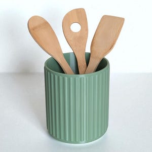 May include: A sage green cylindrical utensil holder with a vertical ribbed texture containing three light brown bamboo kitchen utensils, including a solid spoon, a spoon with a circular hole in the center, and a flat-edged spatula.