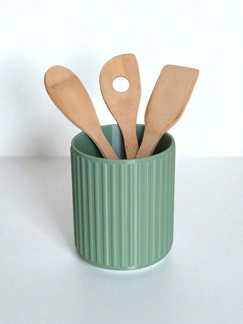 May include: A sage green cylindrical utensil holder with a vertical ribbed texture containing three light brown bamboo kitchen utensils, including a solid spoon, a spoon with a circular hole in the center, and a flat-edged spatula.