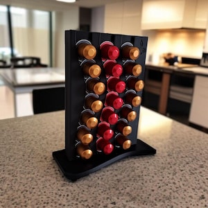 May include: A black vertical coffee pod holder with a wavy design, featuring three columns of slots filled with gold, brown, and red coffee capsules. The stand has a flat base and a small coffee cup icon engraved on the top right corner.