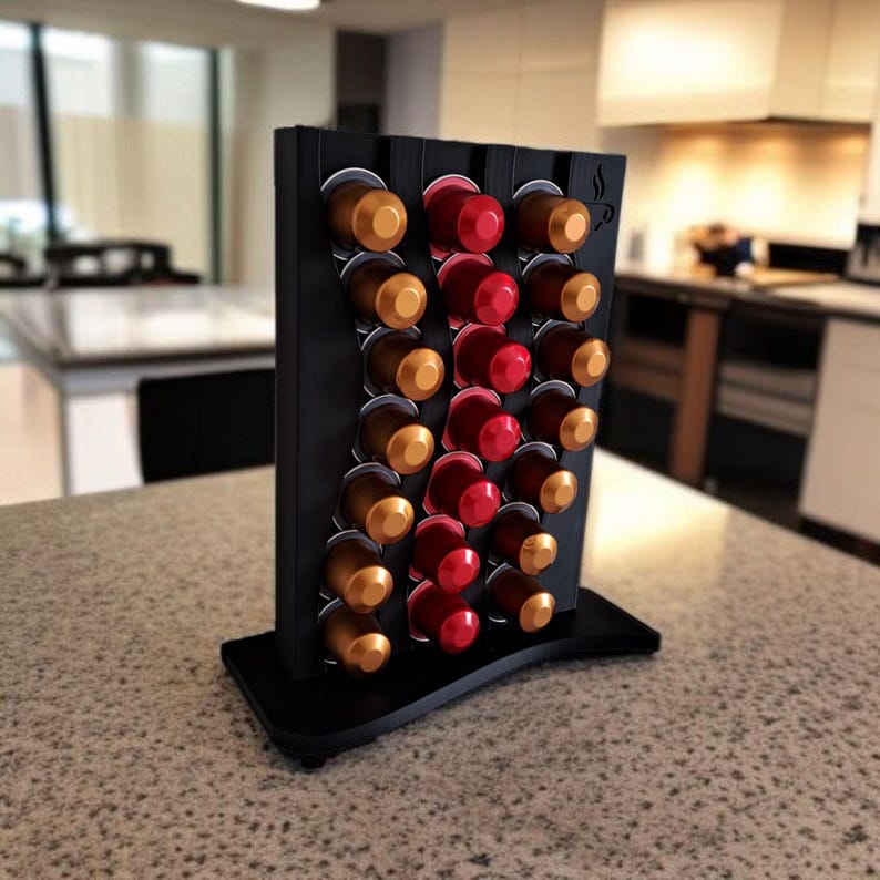 May include: A black vertical coffee pod holder with a wavy design, featuring three columns of slots filled with gold, brown, and red coffee capsules. The stand has a flat base and a small coffee cup icon engraved on the top right corner.