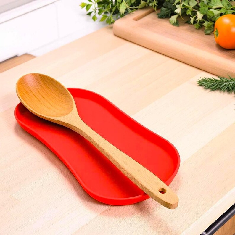 May include: A light brown wooden spoon resting diagonally on a bright red oval silicone spoon rest. The spoon features a smooth, rounded bowl and a long handle with a small circular hole at the end for hanging. The spoon rest has raised edges.
