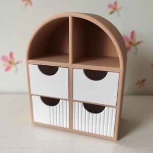 May include: A beige desktop organizer with an arched top, featuring two open upper compartments and four lower drawers. The two bottom drawers have a vertical ribbed texture on their front panels, while all four drawers have semi-circular cutouts.